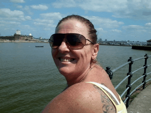 a woman wearing sunglasses and a yellow tank top smiles in front of a body of water