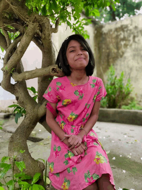 a little girl in a pink floral dress sits under a tree