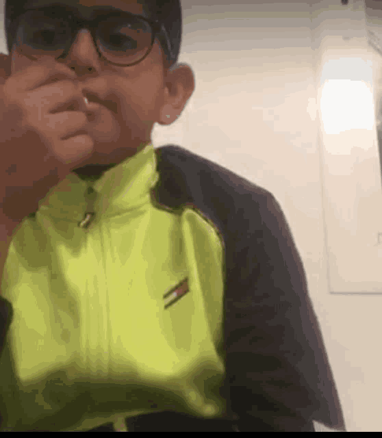 a young boy wearing glasses and a neon yellow jacket is covering his nose .