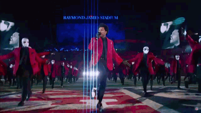 a man in a red jacket stands in front of a crowd of masked dancers at raymond james stadium ..