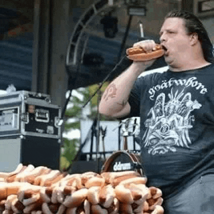 a man is eating a hot dog in front of a microphone while standing in front of a pile of hot dogs .