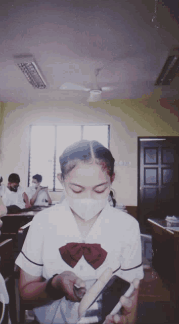 a girl wearing a mask looks at her phone in a classroom with other students