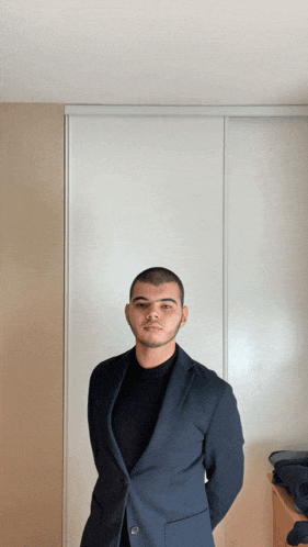 a young man in a suit stands in front of a white wall