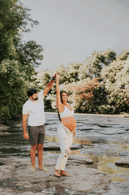 a man and a pregnant woman are dancing on a rock near a river