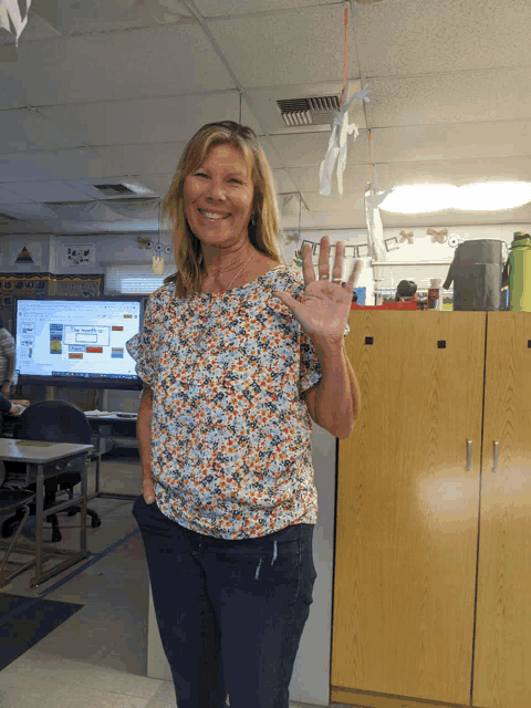 a woman wearing a floral shirt is waving in front of a classroom