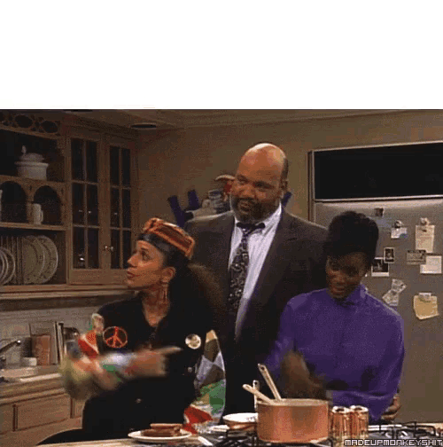 a man in a suit and tie stands between two women in a kitchen with a peace sign on her jacket