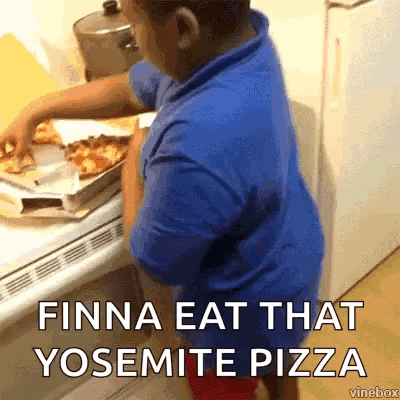 a young boy in a blue shirt is standing in front of a stove taking a pizza out of a box .