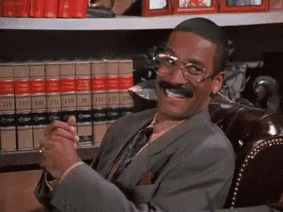 a man in a suit and tie is smiling while sitting in a chair in front of a shelf of books .