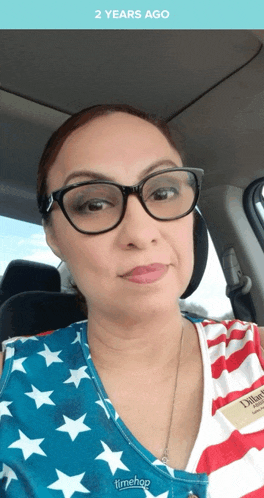 a woman wearing glasses and a timeshop shirt is sitting in a car