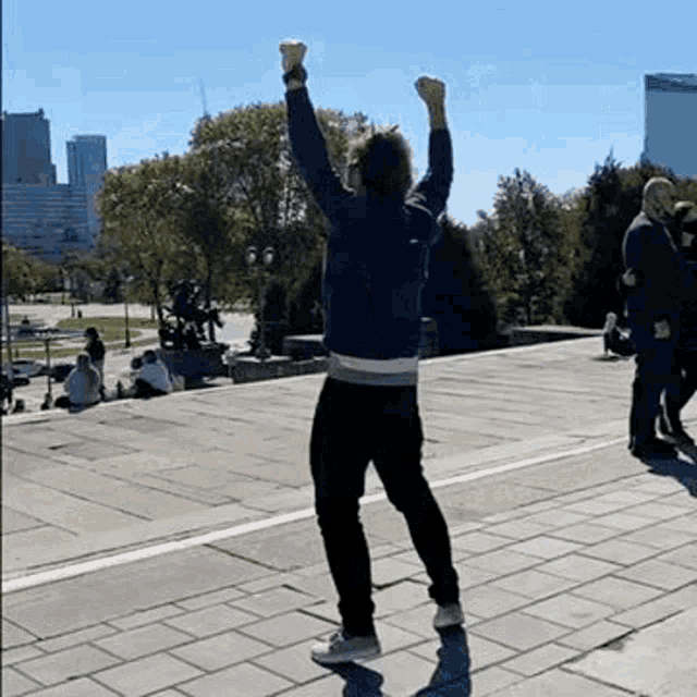 a man is standing on a sidewalk with his arms in the air and people sitting on the steps behind him .