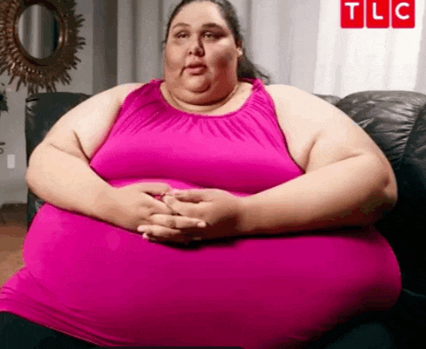 a very fat woman in a pink tank top is sitting on a couch with her hands on her hips .