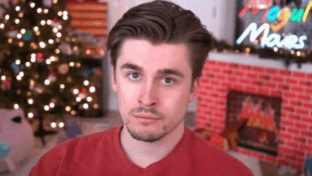 a young man in a red shirt is sitting in front of a christmas tree and fireplace .