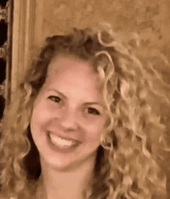 a woman with blonde curly hair is smiling for the camera .