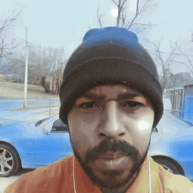 a man with a beard wearing a beanie looks at the camera with a blue car in the background