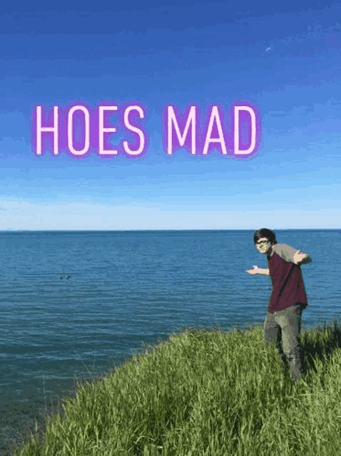 a man is standing on a cliff overlooking the ocean with the words `` hoes mad '' above him .