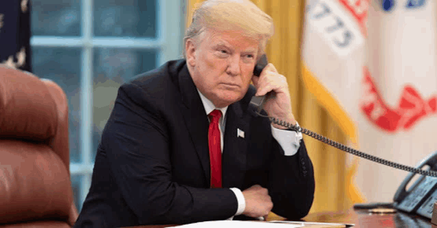 donald trump sits at a desk talking on a phone in front of a flag that says 1776