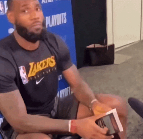 a man wearing a lakers t-shirt sits in front of a blue wall