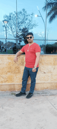 a man wearing sunglasses and a red shirt leans against a stone wall