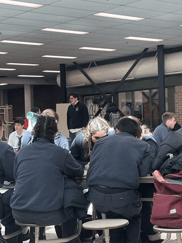 a group of people sit at a table in a cafeteria with a man standing behind them