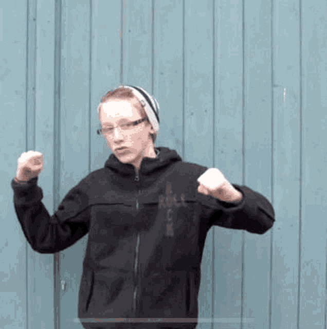 a young man wearing glasses and a hat is standing in front of a blue wooden wall and making a funny face .