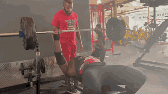 a man lifting a barbell in a gym with a man in a red shirt that says it all on it