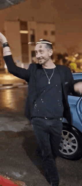 a man wearing a black shirt and a necklace is standing in front of a car