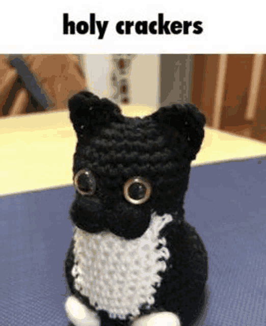 a black and white crocheted cat is sitting on a table with the words `` holy crackers '' above it .