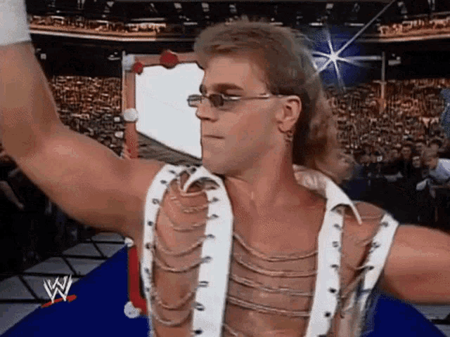 a wrestler wearing sunglasses and a white shirt is standing in front of a crowd and raising his hand in the air .