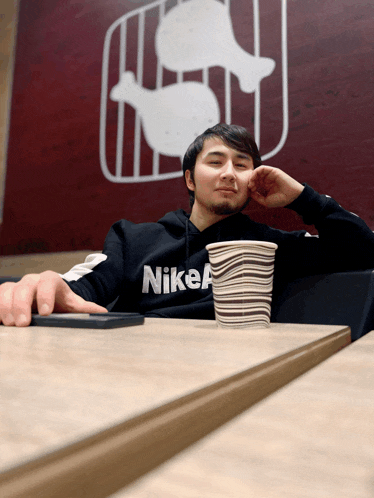 a man wearing a nike hoodie sits at a table with a cup of coffee