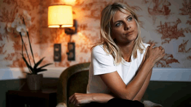 a woman in a white t-shirt sits in a chair in front of a lamp