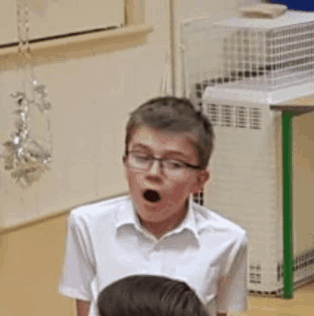 a young boy wearing glasses and a white shirt is singing into a microphone in a classroom .