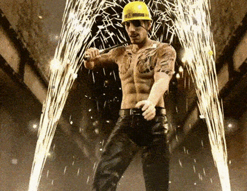 a shirtless man wearing a hard hat and sunglasses stands in front of sparklers