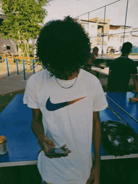 a man with curly hair wearing a white nike t-shirt is holding a cell phone in front of his face .