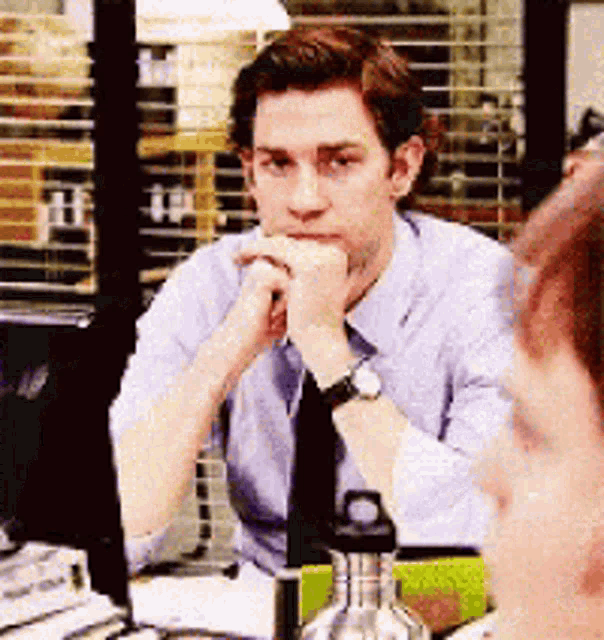 a man in a blue shirt and tie is sitting at a desk with his hand on his chin .