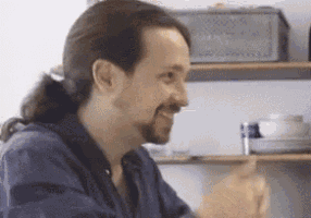 a man with long hair and a beard is smiling while sitting at a table with a shelf in the background .