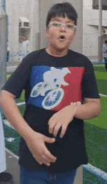 a young boy wearing a black t-shirt with a bicycle on it is standing in front of a soccer field .