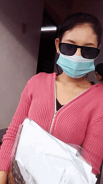 a woman wearing a pink sweater and a mask holds papers