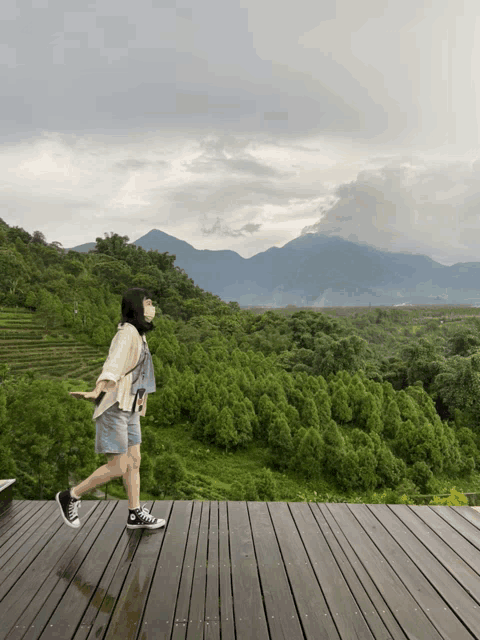 a woman wearing a mask stands on a wooden deck overlooking a lush green forest