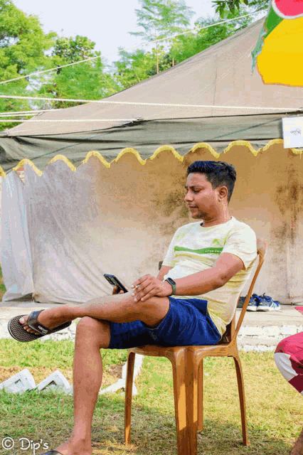 a man sits in a chair with his legs crossed in front of a tent and looks at his phone