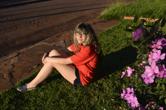 a girl in a red shirt sits in the grass near some purple flowers