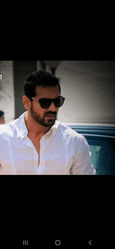 a man wearing sunglasses and a white shirt stands in front of a car .