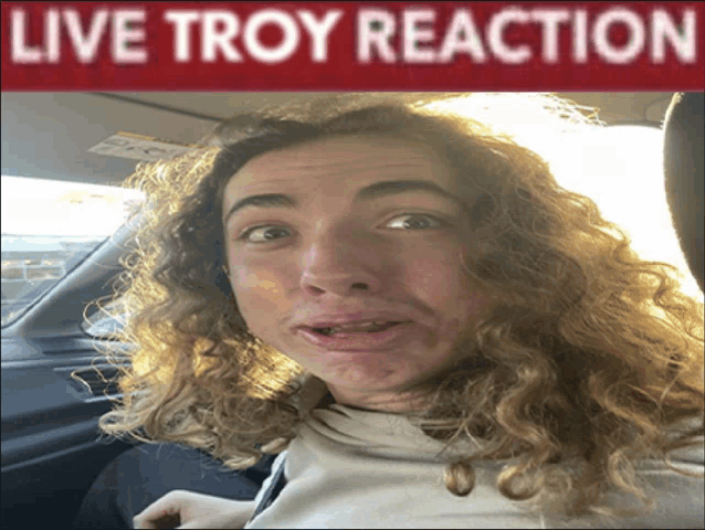 a man with curly hair is sitting in a car under a live troy reaction banner