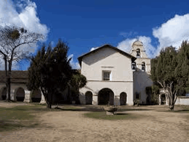 a large white church with a bell tower and arches is surrounded by trees and grass .