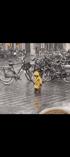 a little boy in a yellow raincoat is standing in the rain in front of a row of bikes .