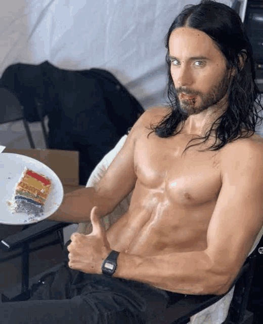 a shirtless man with long hair is sitting in a chair holding a plate of rainbow cake and giving a thumbs up .