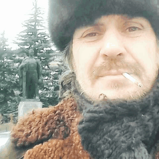 a man in a fur hat and scarf is smoking a cigarette in front of a statue .