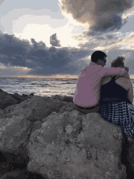 a man in a pink shirt and a woman in a black dress sit on a rock near the ocean