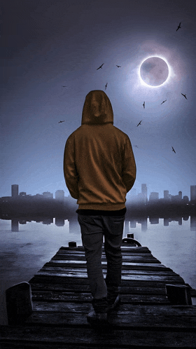 a man in a yellow hoodie is standing on a dock looking at the moon