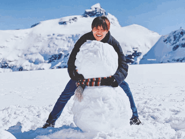 a man is making a snowman in the snow with a mountain in the background and a watermark that says fwdfwdfwd