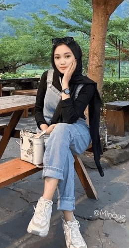 a woman wearing a hijab and overalls is sitting on a wooden picnic table .